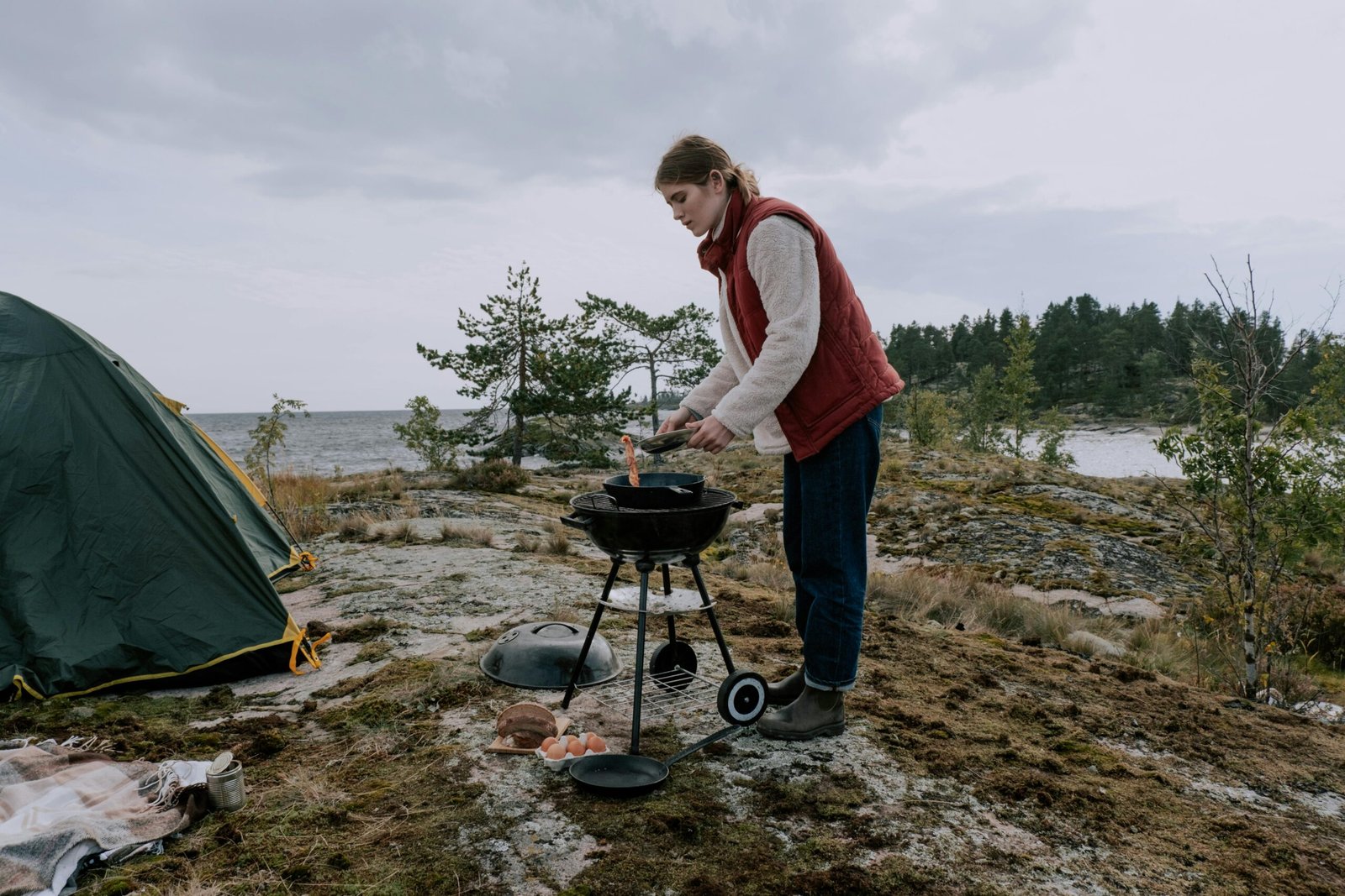 A woman cooks on a portable grill at a scenic seaside campsite.